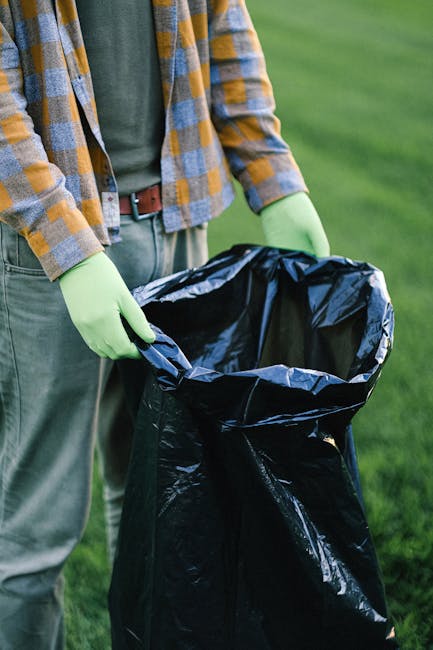 A person wearing white sneakers, navy shorts, and a light blue T-shirt is seen picking up or examining large, irregularly shaped rocks on a rocky shoreline. The rocks vary in size and have a rough, weathered texture with grey, brown, and greenish hues, some covered with patches of moss or algae. The individual is holding a trash picker tool, suggesting they may be collecting debris or cleaning up the area. An empty white trash bag hangs from their arm, indicating an activity related to waste collection or environmental cleanup. The background features more rocks and a natural, coastal environment, with no visible man-made structures. The scene appears well-lit with natural light, emphasizing the varied textures and earthy colors of the stones. Although the setting is outdoors rather than within a waste management context, this scene highlights the importance of responsible waste disposal and environmental preservation, themes that align naturally with services such as rubbish removal or on-site clearance offered by companies like Rubbish Removal Fulham.