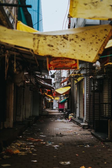 A narrow, deserted alleyway with a rough, uneven pavement surface littered with various bits of rubbish and debris. The sides of the alley are lined with closed shopfronts, some with metal security shutters, and a few with partially visible signs. Overhead, several makeshift awnings and panels, made from worn, weathered materials such as wood and fabric, hang at irregular angles, with visible dirt and stains. The lighting appears subdued, with natural daylight filtering through gaps and crevices, casting soft shadows along the ground. In the background, a few structures and antennas are faintly visible, emphasizing an urban environment. The scene suggests a location that might benefit from private waste collection or rubbish removal services, with Rubbish Removal Fulham specializing in on-site clearance for commercial properties along North End Road. The overall atmosphere is slightly cluttered and neglected, highlighting the need for professional waste management to maintain cleanliness and sanitation in this densely built area.