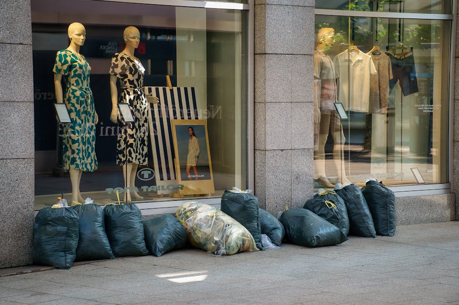 A collection of dark green garbage bags, some tightly sealed and others partially open revealing mixed waste inside, lined up on the sidewalk in front of a clothing store window on a city street. The store's glass facade displays mannequins dressed in patterned dresses and casual shirts with trousers, along with framed photographs of models wearing the clothing. The bags are placed directly on the paved surface, with a few leaning against the store's exterior wall. The scene appears to indicate a private waste collection or rubbish removal activity, supported by the presence of the bags rather than communal or public trash bins, aligning with services like those offered by Rubbish Removal Fulham for commercial waste management. Bright natural daylight illuminates the area, casting shadows on the pavement and highlighting the textures of the plastic bags and the store's glass and stone materials.