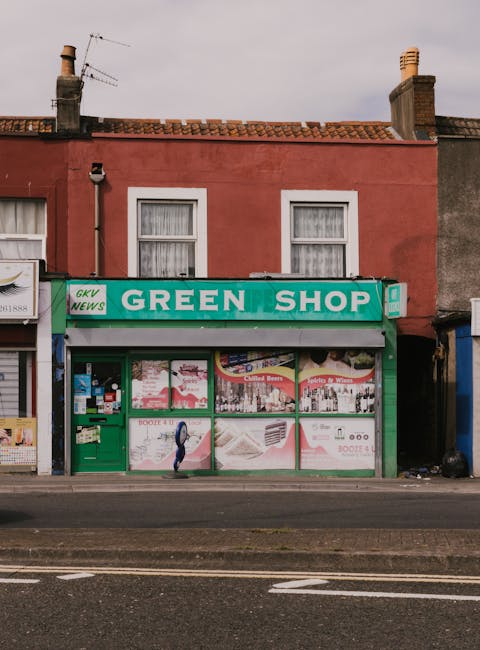 A narrow, deserted alleyway with a rough, uneven pavement surface littered with various bits of rubbish and debris. The sides of the alley are lined with closed shopfronts, some with metal security shutters, and a few with partially visible signs. Overhead, several makeshift awnings and panels, made from worn, weathered materials such as wood and fabric, hang at irregular angles, with visible dirt and stains. The lighting appears subdued, with natural daylight filtering through gaps and crevices, casting soft shadows along the ground. In the background, a few structures and antennas are faintly visible, emphasizing an urban environment. The scene suggests a location that might benefit from private waste collection or rubbish removal services, with Rubbish Removal Fulham specializing in on-site clearance for commercial properties along North End Road. The overall atmosphere is slightly cluttered and neglected, highlighting the need for professional waste management to maintain cleanliness and sanitation in this densely built area.