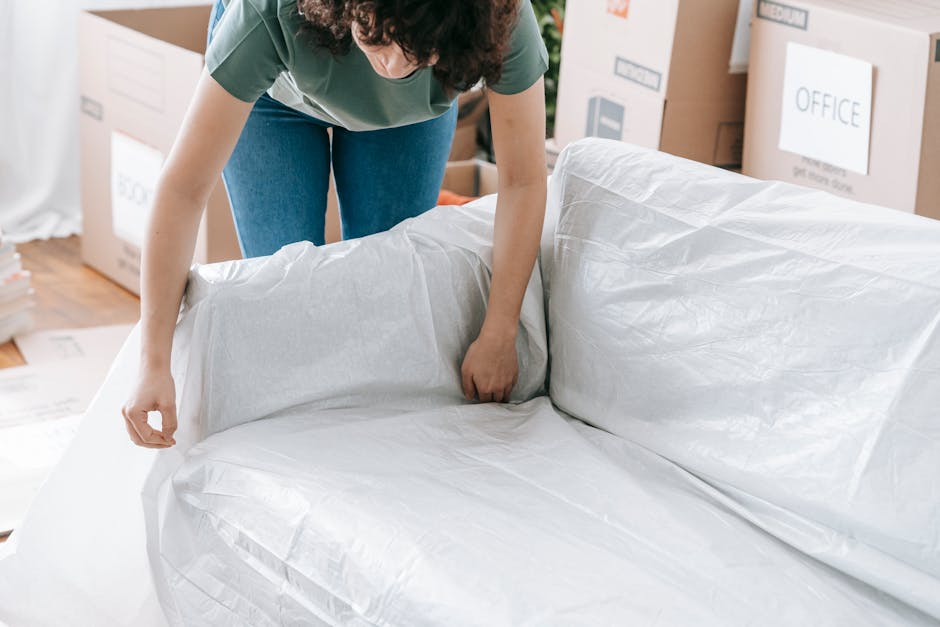 A person with curly hair, wearing a green t-shirt and blue jeans, is in the process of wrapping a large mattress with white plastic protection. The mattress appears thick with a light-colored fabric surface, possibly grey or beige, and is being securely covered for removal or disposal. The scene is set indoors, likely in a home or storage area, with cardboard boxes and packaging materials visible in the background, some labeled with signs like 'OFFICE' and 'MEDIA'. The person is standing on a wooden floor, and the environment around them is filled with packing supplies indicative of a move or clearance process, which may relate to independent waste collection or private rubbish removal services like those offered by Rubbish Removal Fulham. Proper packaging methods are evident, aligning with professional rubbish or waste clearance practices to ensure safe handling of large items such as mattresses during the disposal process.