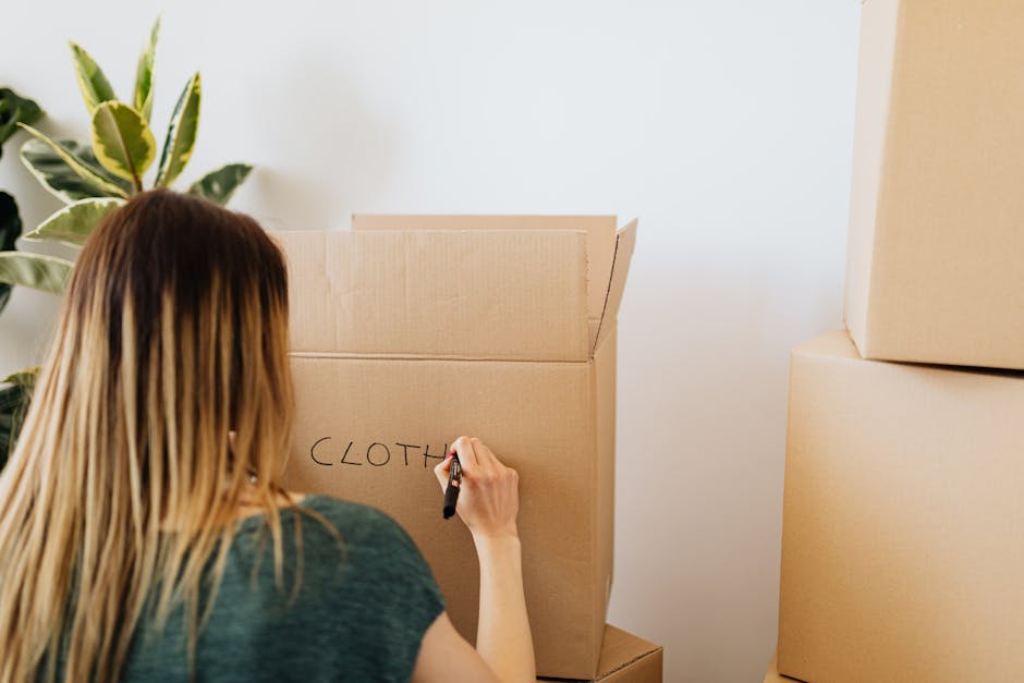 A woman with long, straight, light brown hair, viewed from behind, is writing the word 'CLOTH' with a black marker on the side of a large, open cardboard box. The box appears to be made of standard corrugated material with a light brown, natural finish. It is positioned indoors, likely in a domestic setting, with a white wall in the background. To the left of the woman, a potted plant with broad, variegated green and cream leaves is partially visible, adding a natural element to the scene. Adjacent to the open box, there are several other cardboard boxes stacked or placed nearby, suggesting an organized process for packing or moving. The lighting is soft and natural, illuminating the scene evenly, indicating ambient daylight. This context aligns with the activities of clearing or preparing waste materials, typical in private rubbish removal or on-site clearance services, subtly reflecting the theme of alternative waste handling options. Rubbish Removal Fulham may be involved in the process, given the location and service relevance.