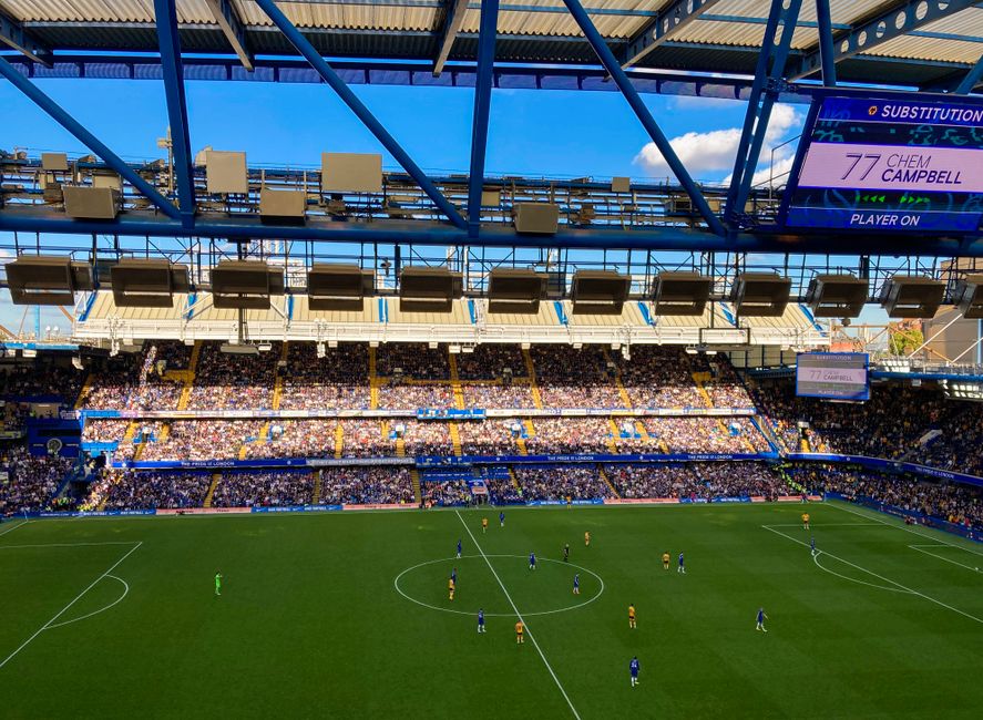 Inside a large football stadium with a partially open roof, the image shows a view of the pitch from behind the stands. The playing field is green with clearly visible white boundary lines, and players are positioned across the field, some near the center circle, dressed in blue and yellow kits. The stadium seating is filled with spectators, creating a vibrant, bustling atmosphere. Above the field, a digital scoreboard displays the text '77 CHEM CAMPBELL' and indicates the match is ongoing. Overhead, numerous lighting fixtures hang from the blue steel framework of the roof structure, which contrasts with the partly cloudy sky visible through the open sections. The environment is bright, with natural sunlight illuminating the seating areas and the pitch. The scene could relate to arrangements for match day events, possibly involving minor clearing or maintenance activities by services like Rubbish Removal Fulham, working in the context of event waste management beyond just standard rubbish collection in surrounding areas.